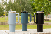 Load image into Gallery viewer, Three insulated travel mugs in blue, green, and black on a stone surface with a blurred natural background.
